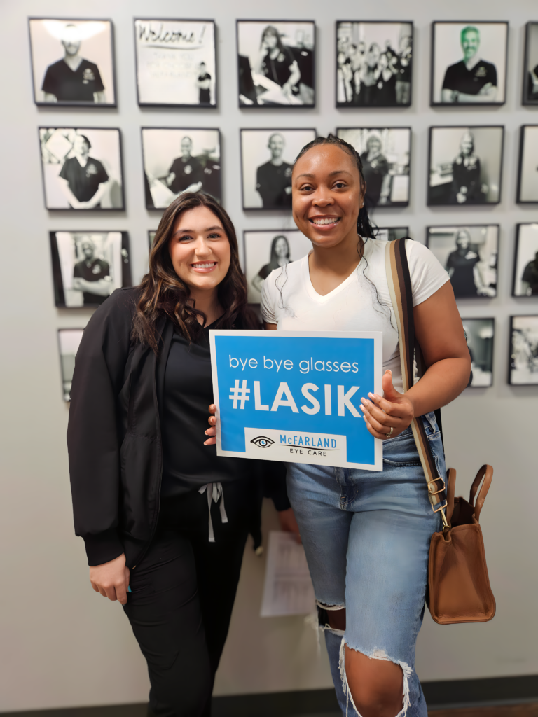 Two smiling women pose together at an eye care clinic while holding a “bye bye glasses #LASIK” sign after a LASIK consultation or procedure.
