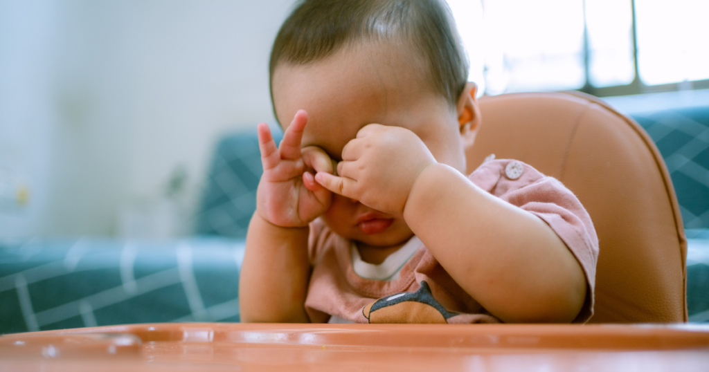 Young child rubbing both eyes while sitting in a high chair.
