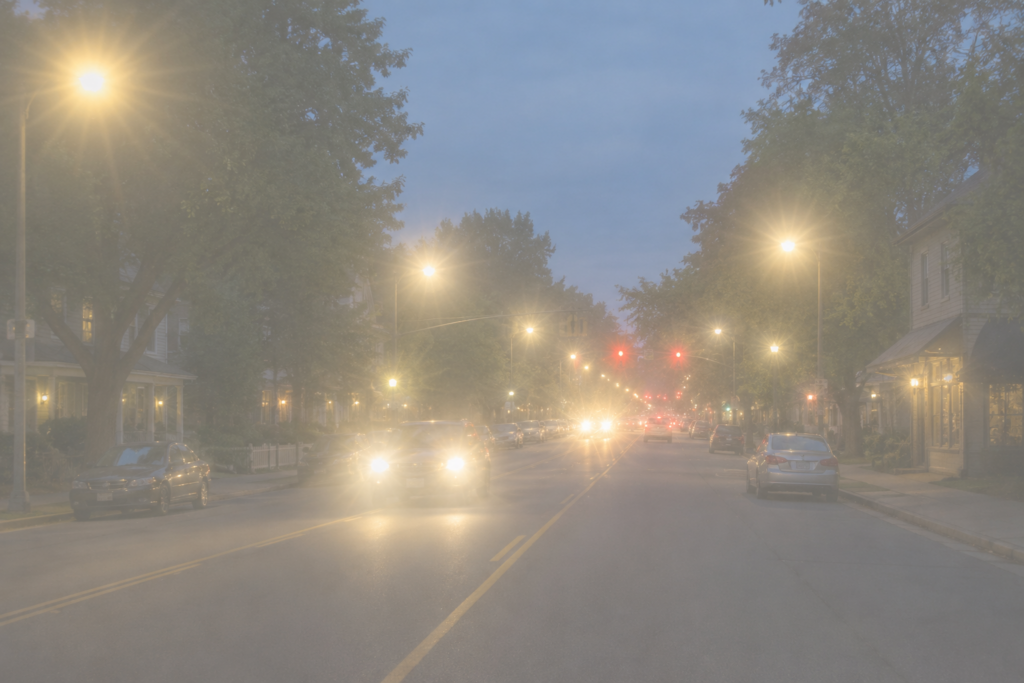 Simulation of cloudy vision showing a hazy street at dusk with muted colors, glare and bright halos around headlights and streetlights.